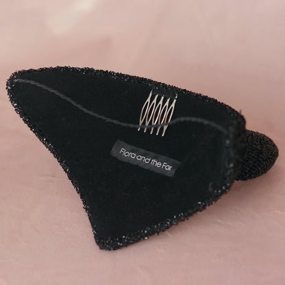 A black devil hat lined in velvet with a hat elastic and a metal comb featuring a fabric label that reads 'Flora and the Fox', set against a soft pink background.