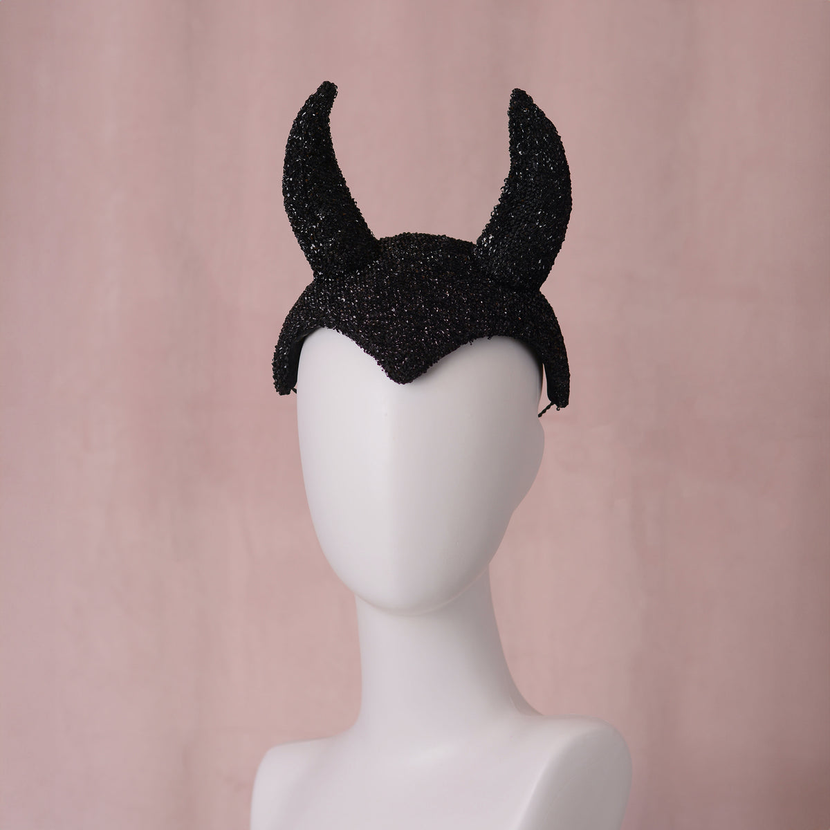 A black glittery devil horn halloween headband displayed on a white mannequin head against a soft pink background.