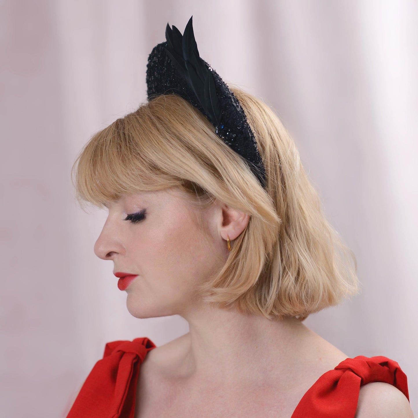 A blonde woman in profile wearing an elegant black funeral fascinator headband and a red dress against a soft pink background.