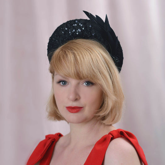 A blonde woman  wearing a black feathered headband style fascinator hat with sequin lace and beading paired with a red dress, photographed against a soft pink curtain background.
