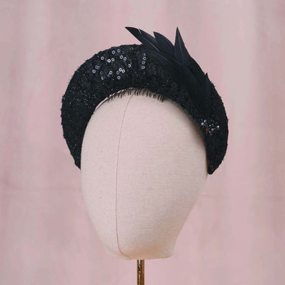 A black evening-wear feathered fascinator hat with sequined detailing and feathers, displayed on a cream fabric mannequin head against a soft pink background.