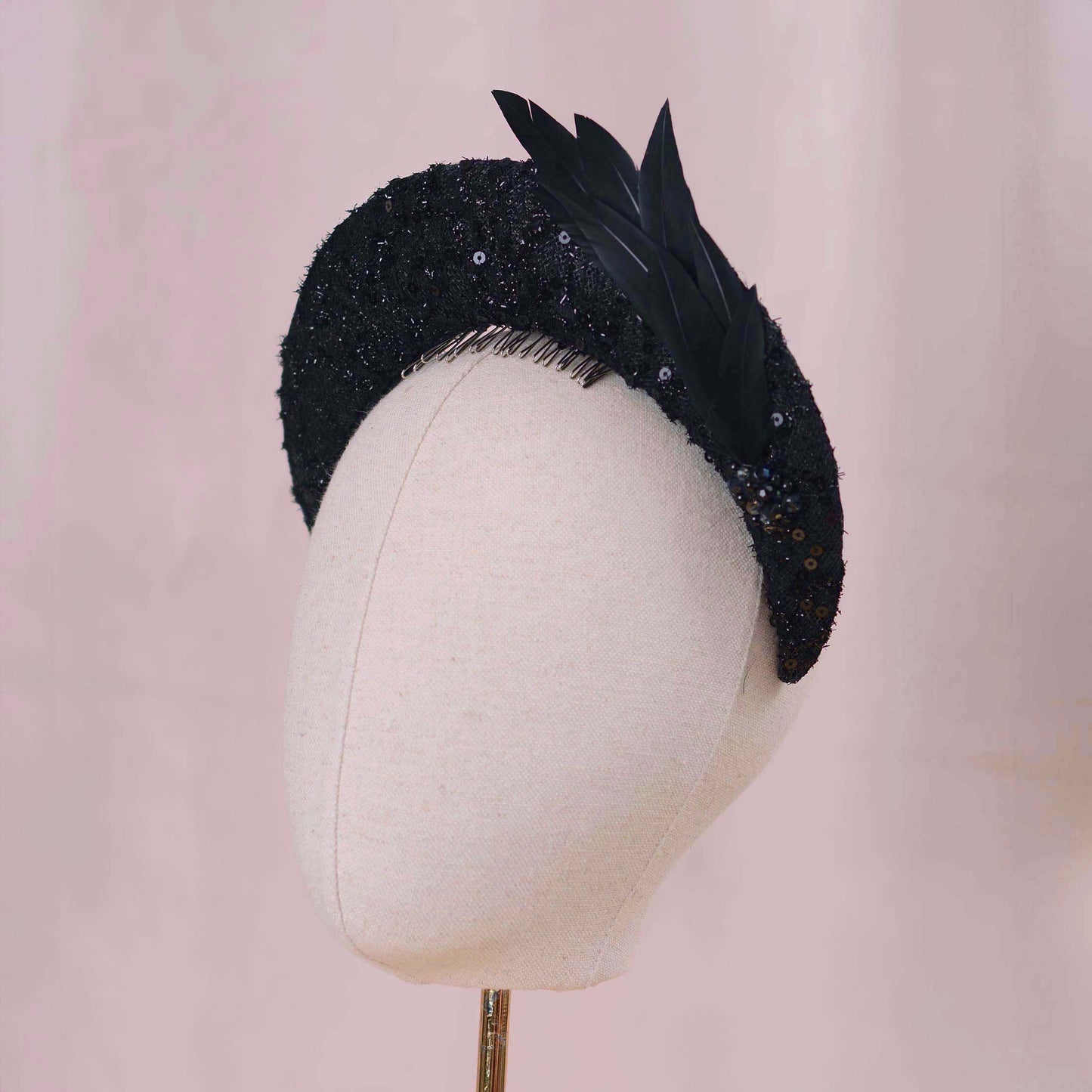 A black evening-wear feathered fascinator hat with sequined detailing and feathers, displayed on a cream fabric mannequin head against a soft pink background.