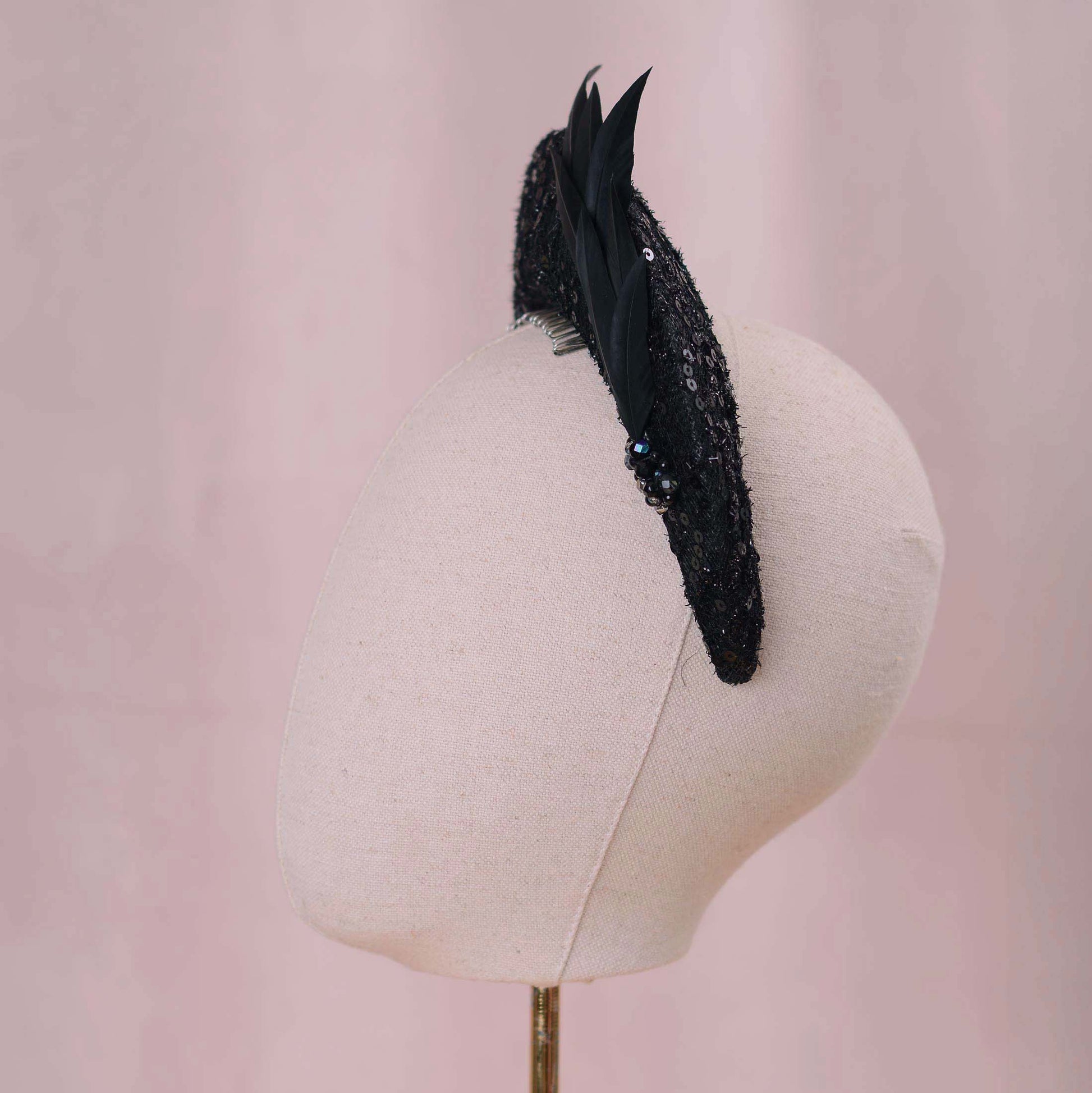 A black feather fascinator with sequined detailing and small black-blue rhinestone accents, displayed on a cream fabric mannequin head against a soft pink background.