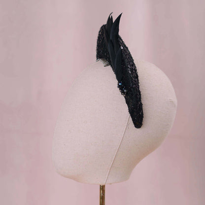 A black feather fascinator with sequined detailing and small black-blue rhinestone accents, displayed on a cream fabric mannequin head against a soft pink background.