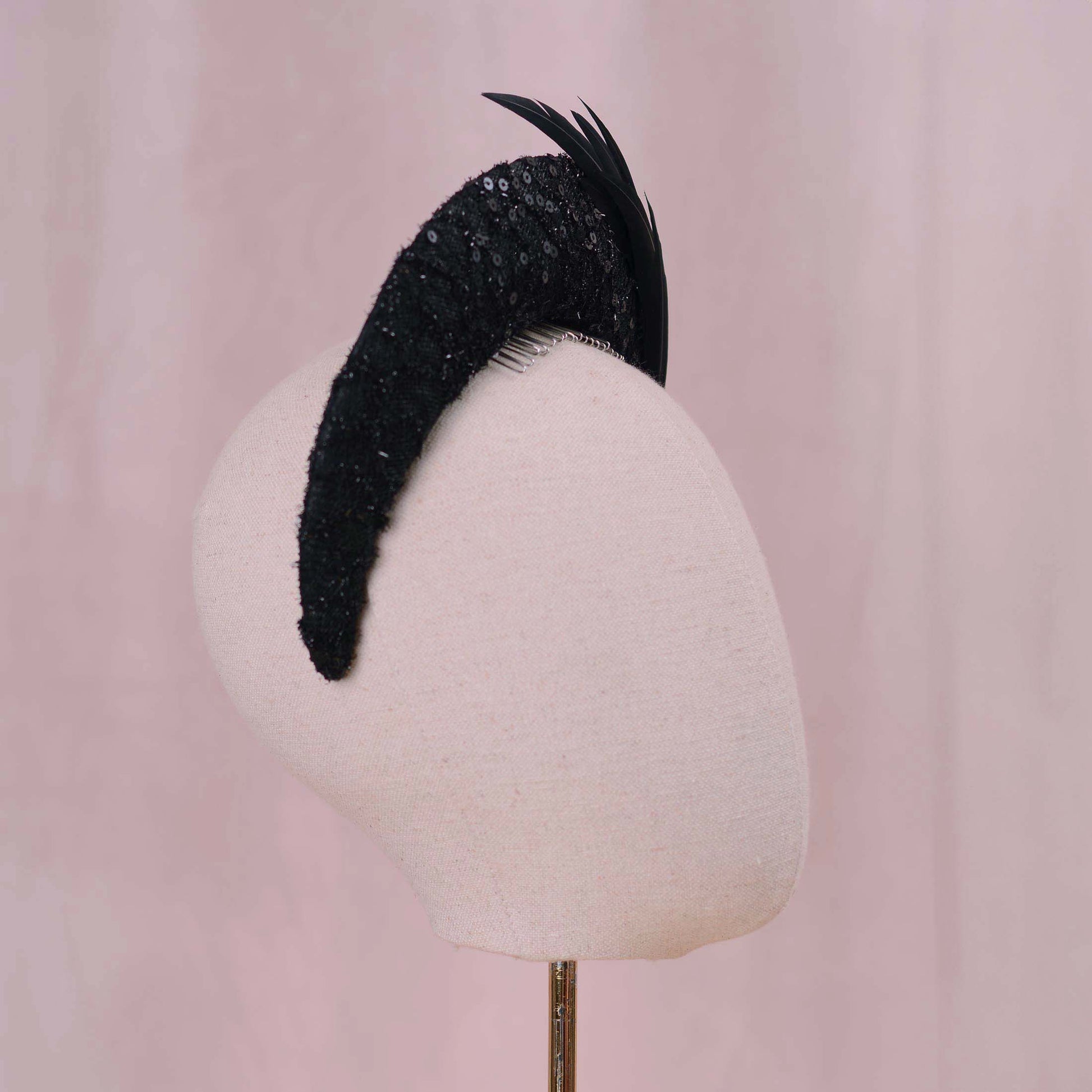 A black feathered fascinator hat with a lace, sequined crescent moon headband base, displayed on a cream-colored mannequin head against a soft pink background.
