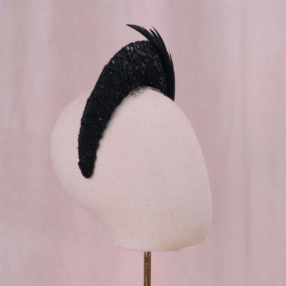 A black feathered fascinator hat with a lace, sequined crescent moon headband base, displayed on a cream-colored mannequin head against a soft pink background.