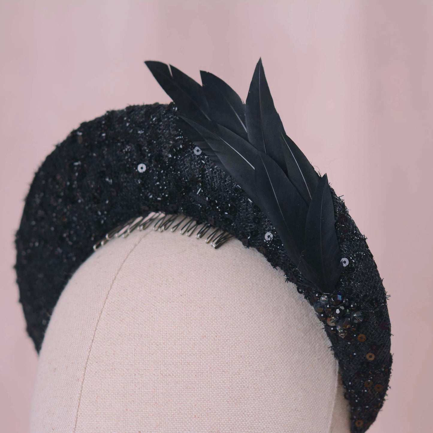 A close-up of a black embellished fascinator headpiece with black sequin lace, feathers and beading displayed on a beige fabric mannequin head against a soft pink background.