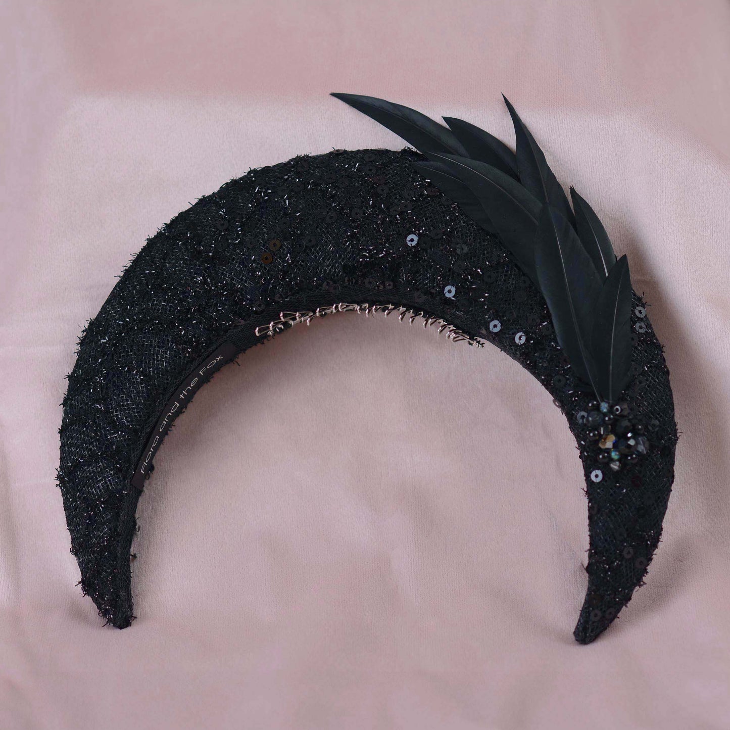 A close-up of a black embellished fascinator headpiece with black sequin lace, feathers and beading displayed on a beige fabric mannequin head against a soft pink background.