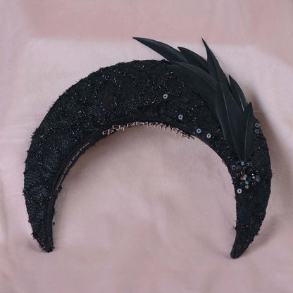 A close-up of a black embellished fascinator headpiece with black sequin lace, feathers and beading displayed on a beige fabric mannequin head against a soft pink background.