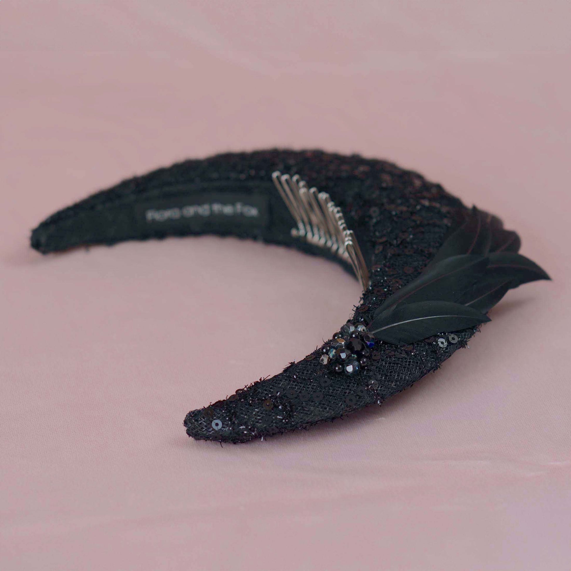 A black sequined crescent moon headband with beading sequins and black feathers, displayed on a soft pink background. The inner band shows  a silver metal comb fitting and the brand name 'Flora and the Fox'.