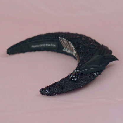 A black sequined crescent moon headband with beading sequins and black feathers, displayed on a soft pink background. The inner band shows  a silver metal comb fitting and the brand name 'Flora and the Fox'.