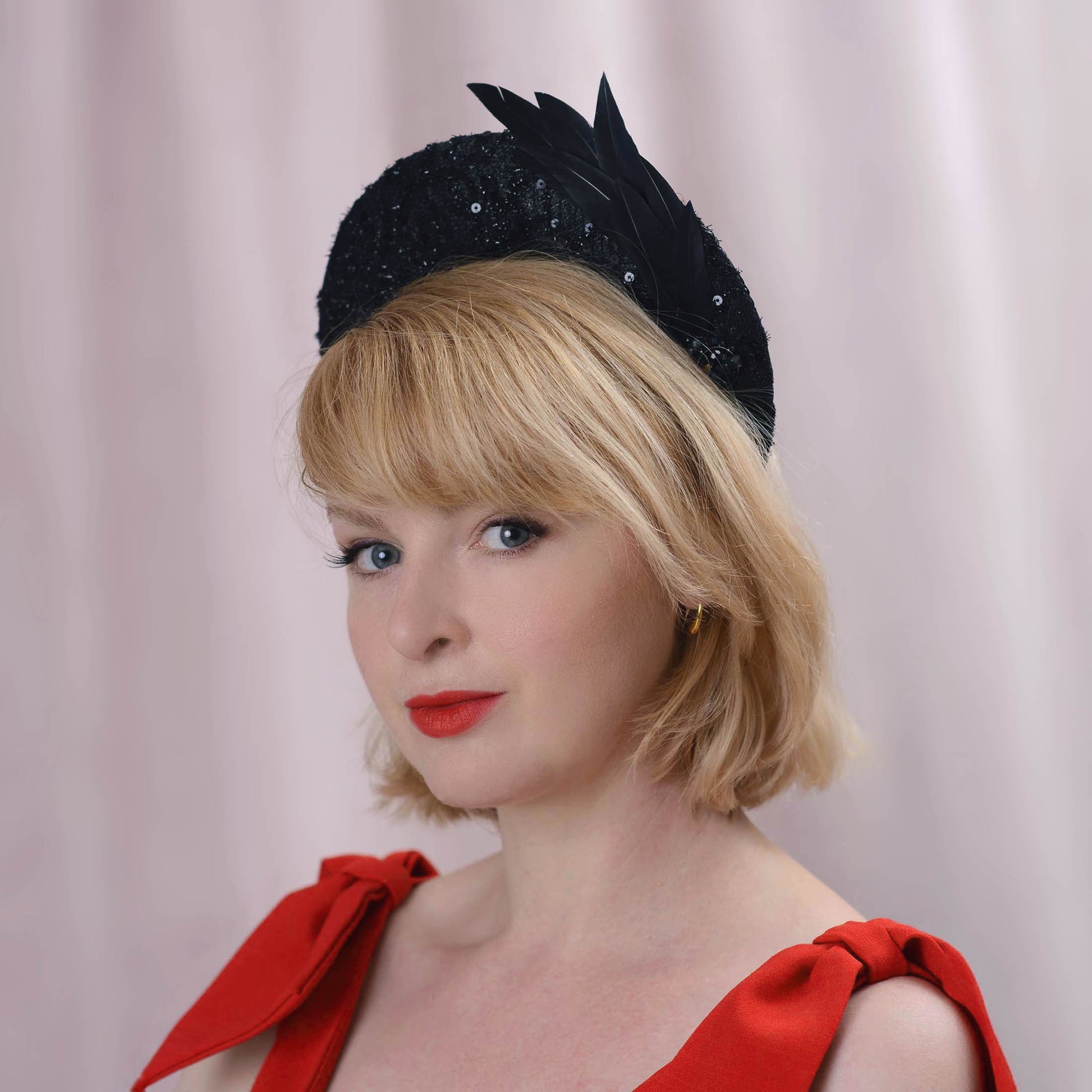 A blonde woman with a short bob wears a black feathered wedding fascinator hat in a tall headband style. It is decorated with lace fabric, sequins, beading and a feather mount. She is wearing a red bow-shoulder dress, posed against a soft pink curtain background.