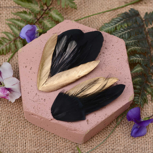 Two decorative feather hair pieces with black and gold feathers arranged on a pink hexagonal stone slab, surrounded by green fern leaves and small purple flowers on a burlap background.
