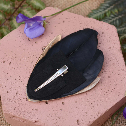 A black feather hair clip with a silver metal alligator-style clip and felt back, displayed on a pink textured stone with a purple flower in the background.