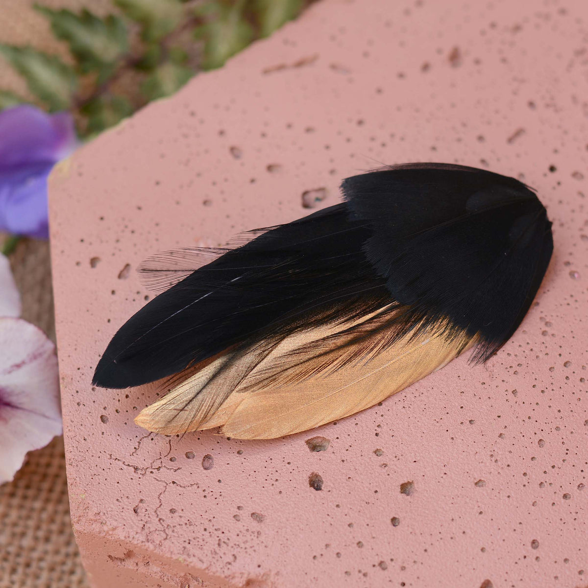 A decorative feathers fascinator with a black and metallic gold feathers, displayed on a textured pink stone surface with blurred purple flowers in the background.