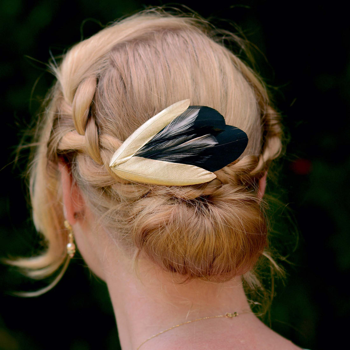 Blonde woman wih a braided bun hair style decorated with a black and gold feather fascinator  in a 190s style. Perfect for weddings and black tie events.