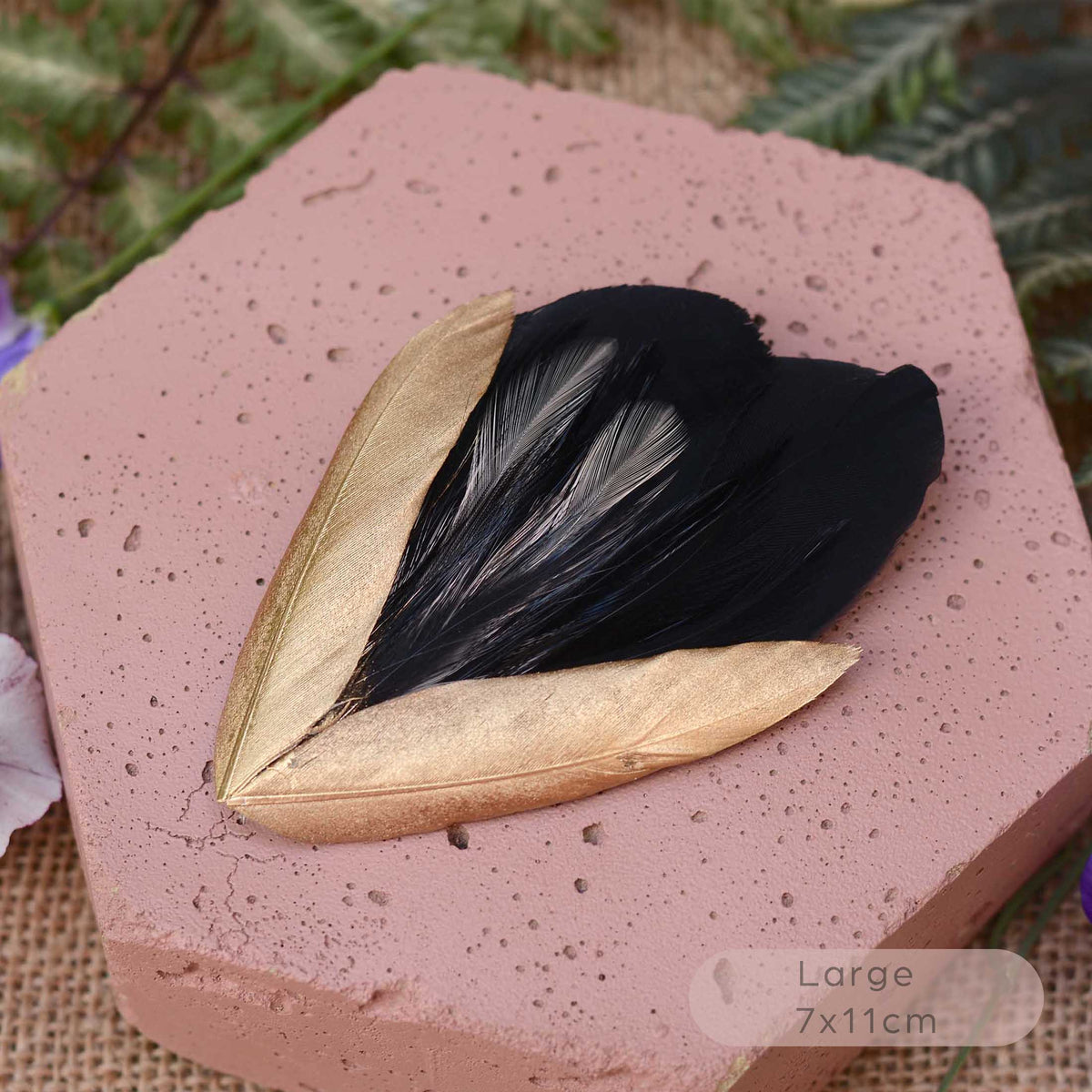 A decorative feather hair ornament with two gold-toned feathers and a black feathers in the center, displayed on a pink hexagonal stone with green foliage in the background.