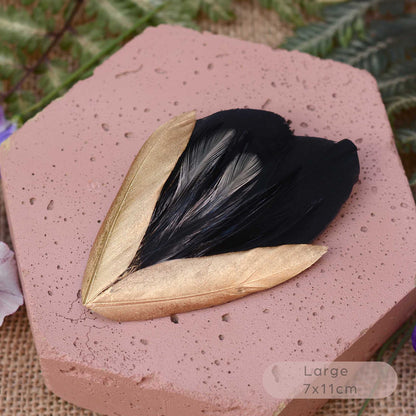 A decorative feather hair ornament with two gold-toned feathers and a black feathers in the center, displayed on a pink hexagonal stone with green foliage in the background.
