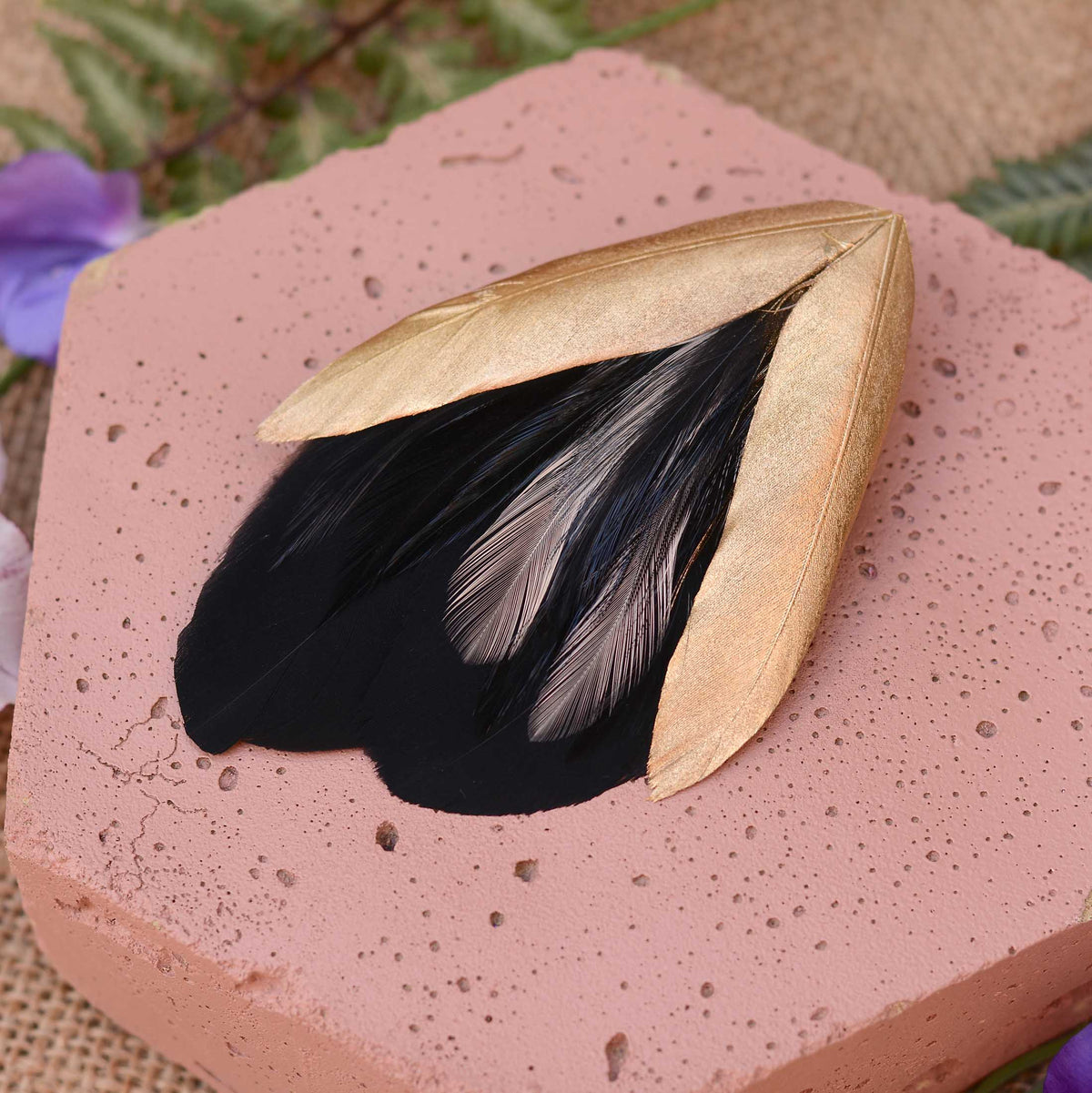A decorative feather fascinator with two gold-toned feathers in a V-shape and black feathers with a hint of blush pink, displayed on a pink stone slab with blurred greenery and flowers in the background.