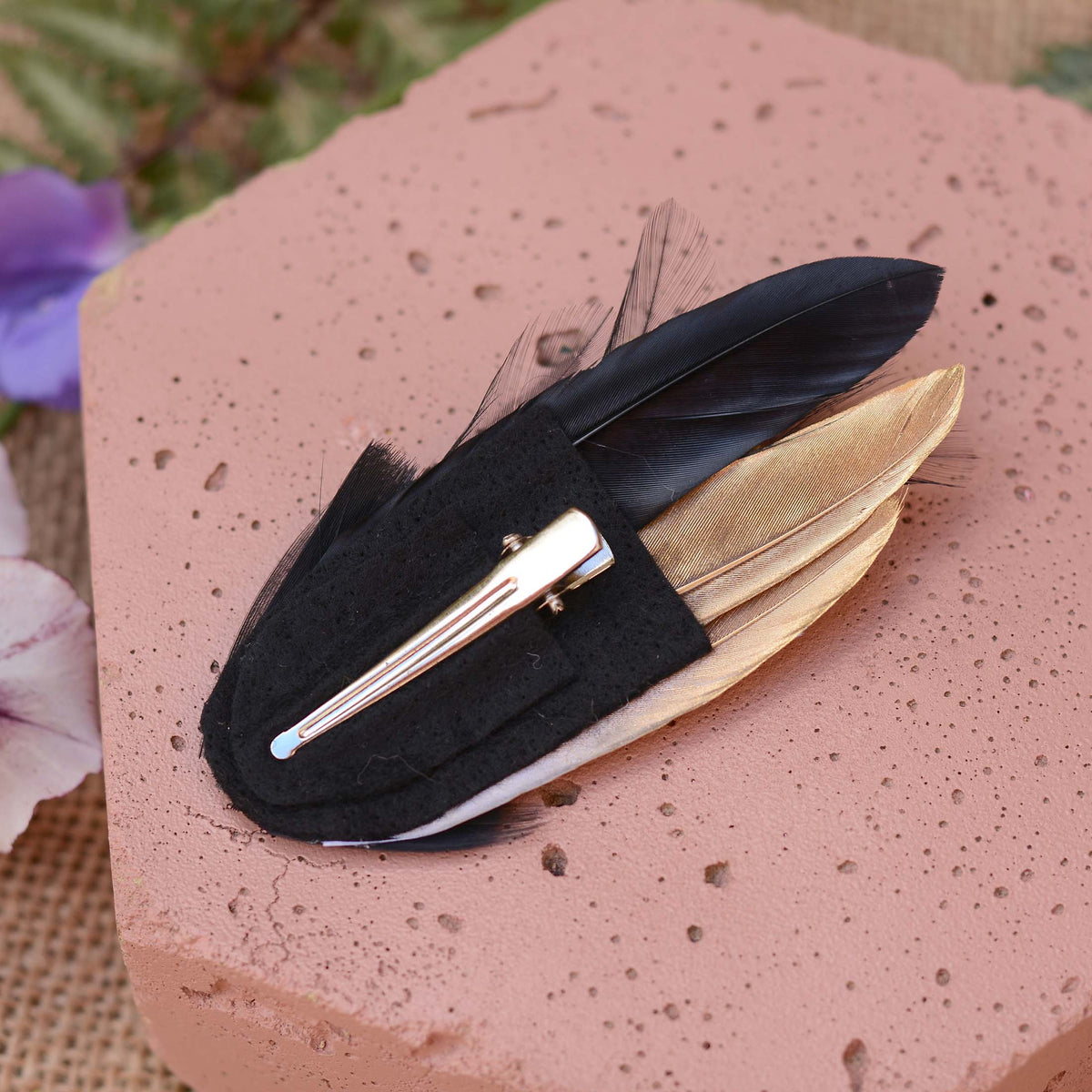 A black feather hair clip with a silver-tone metal alligator-style clip, featuring black and gold feathers, displayed on a terracotta-colored stone block.