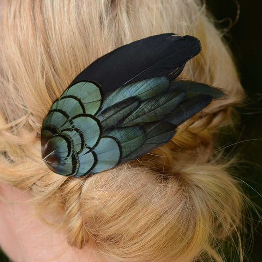 A close-up of a decorative feather hair accessory clipped onto blonde hair. The feather has a black with iridescent blue-green sheen creating a scallop pattern and designed in a wing-like shape.