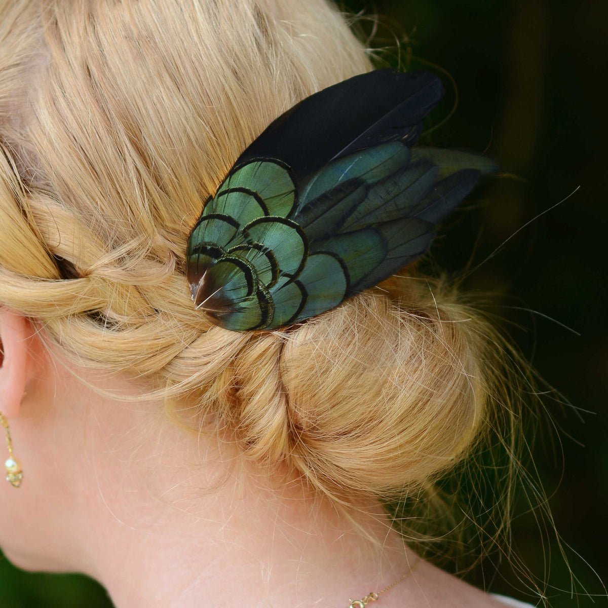 A close-up lifestyle shot of a woman with blonde hair in a twisted bun, wearing a decorative hair accessory made of iridescent green and black feathers.