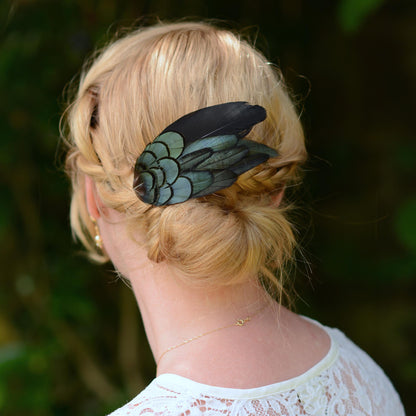 A blonde woman shown from the back wearing a decorative feather hair accessory in her hair. The accessory features a black goose feather layered with black - green irridescent rooster feather against a blurred green background