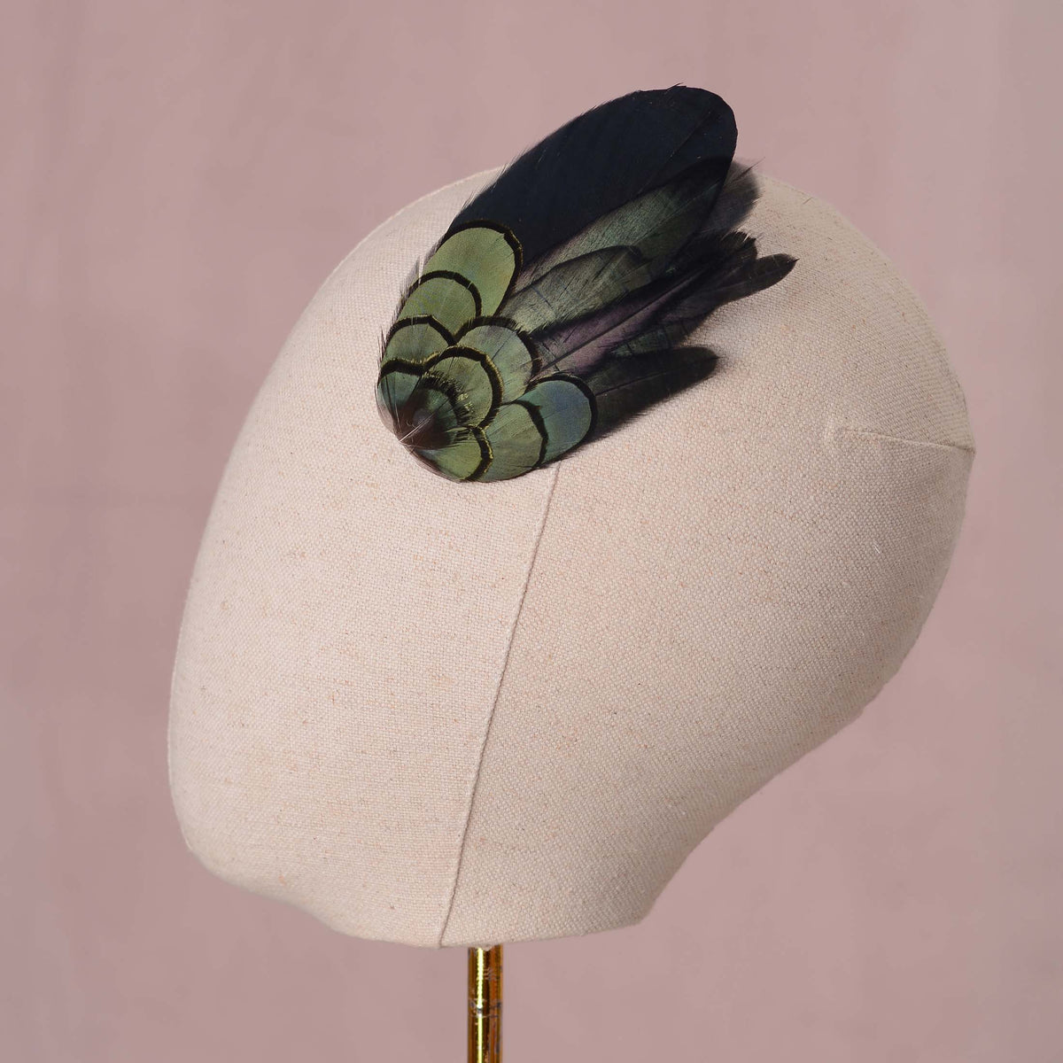 A decorative feather hair accessory with iridescent green and black feathers, displayed on a cream fabric mannequin head against a soft pink background.