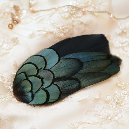 A decorative feather accessory for evening and black tie events with iridescent blue-green and black feathers, laid on a cream lace fabric background with a delicate necklace nearby. Perfect for dark romance and gothic weddings.