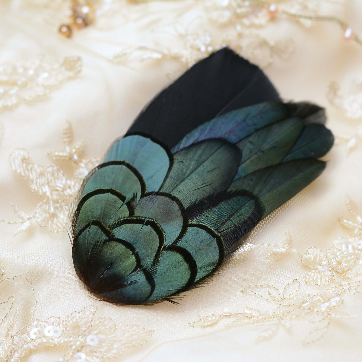 A decorative feather fascinator or hair accessory with iridescent teal, green and black feathers, displayed on a cream lace fabric background.