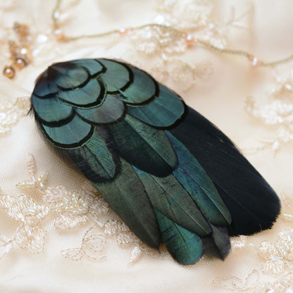 A wing shaped decorative feather ornament with iridescent teal-green and black feathers, laid on a cream lace bridal fabric background with pearl strands.