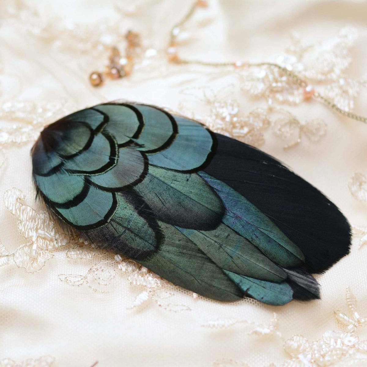 A decorative feather hair barrette with iridescent teal-green and black feathers, placed on a cream lace bridal fabric background with small jewellery props.