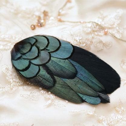 A decorative feather hair barrette with iridescent teal-green and black feathers, placed on a cream lace bridal fabric background with small jewellery props.