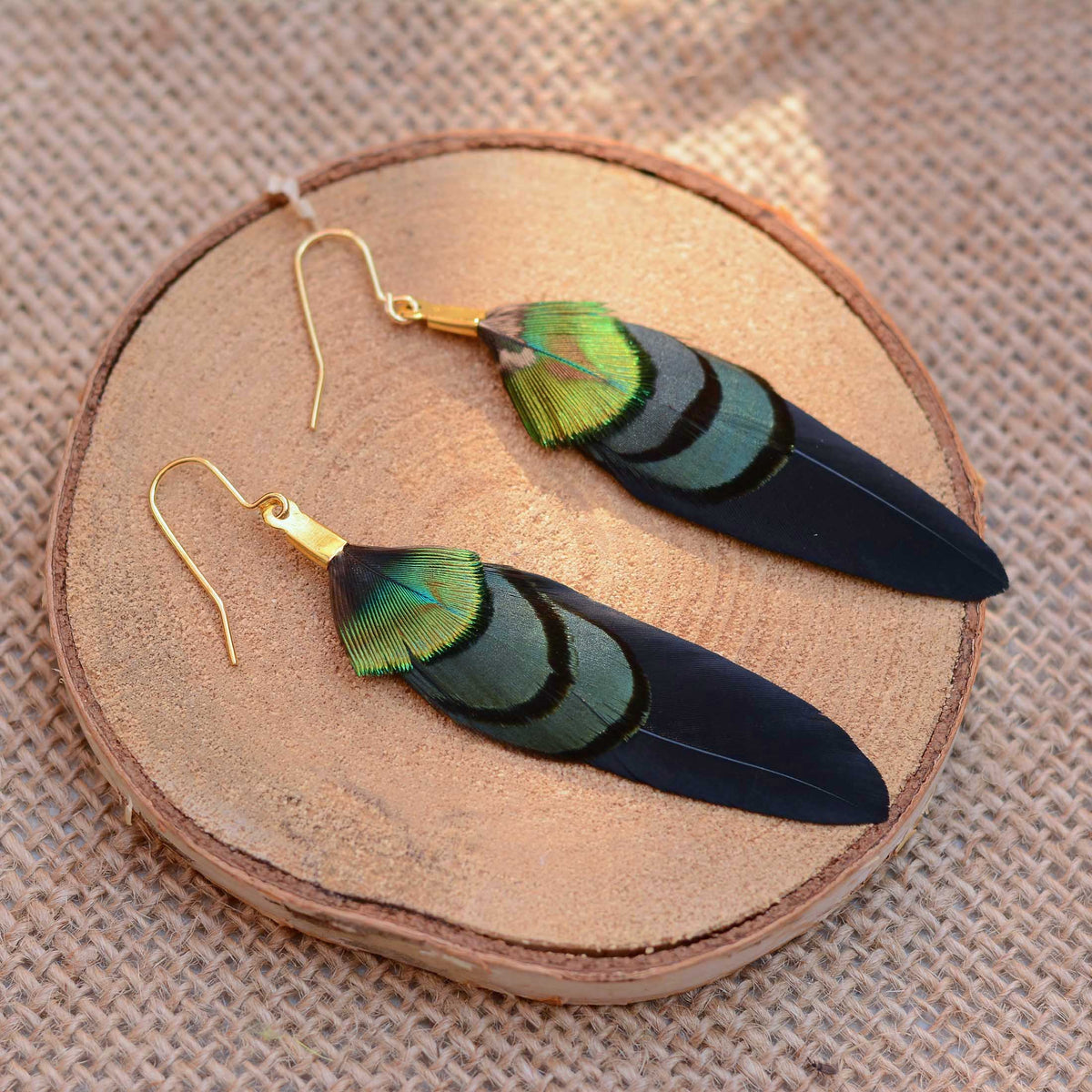 A pair of dangling fstival feather earrings with iridescent green and blue feathers on black wing feathers and gold fishhook wires, displayed on a round wood slice over a textured wood surface.