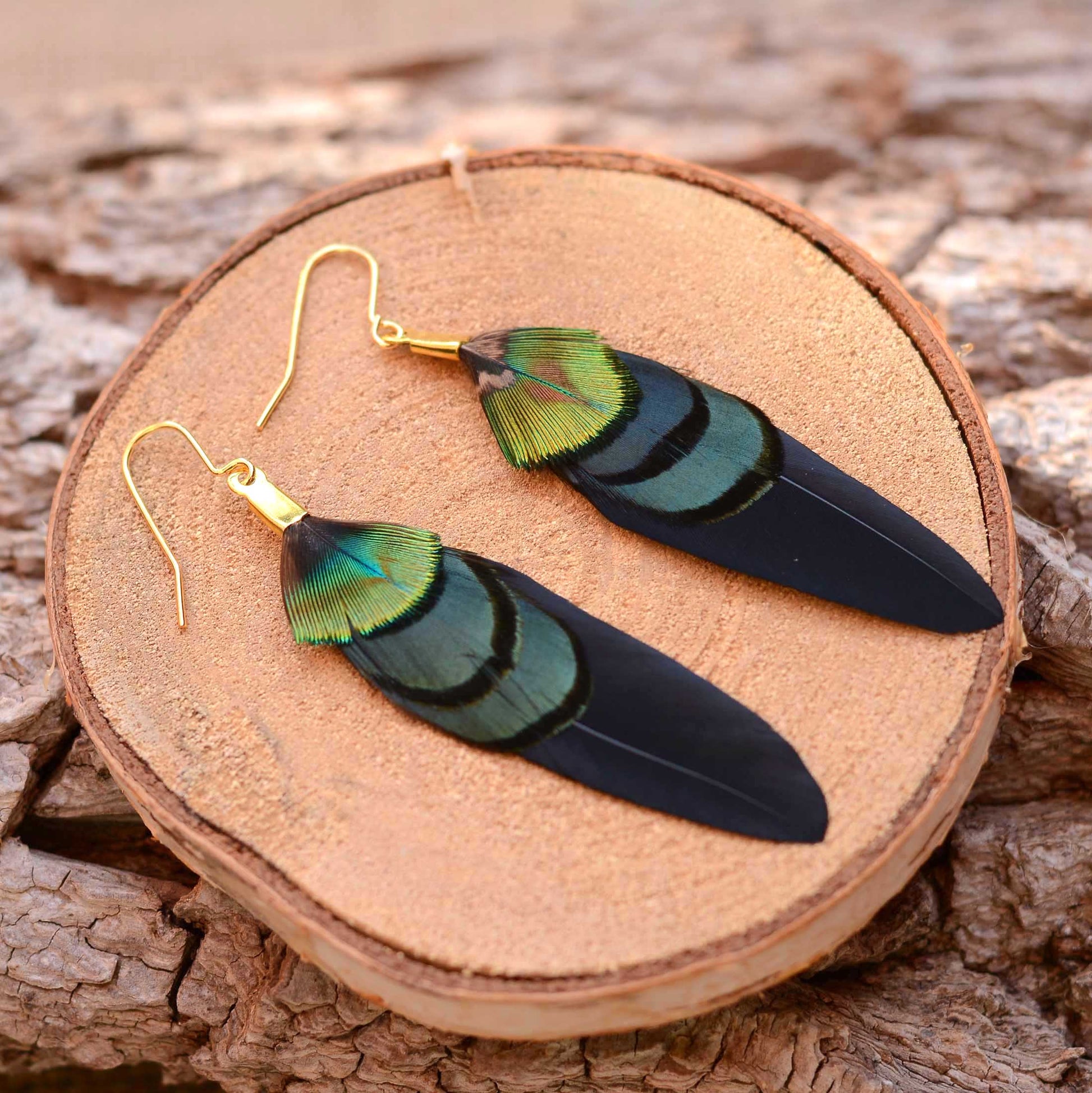A pair of dangling boho feather earrings with iridescent green and blue feathers on black wing feathers and gold fishhook wires, displayed on a round wood slice over a textured wood surface.