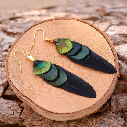 A pair of dangling boho feather earrings with iridescent green and blue feathers on black wing feathers and gold fishhook wires, displayed on a round wood slice over a textured wood surface.