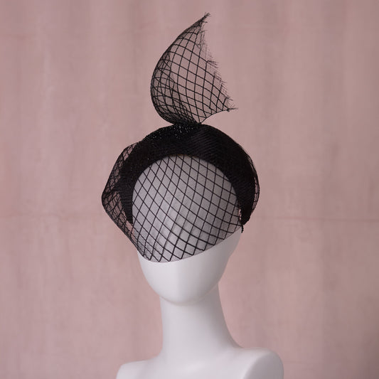 A black fascinator hat with a sculpted mesh veil with a diamond print that sweeps around the face with a tall curved tail above the head, displayed on a white mannequin head against a soft pink backdrop.