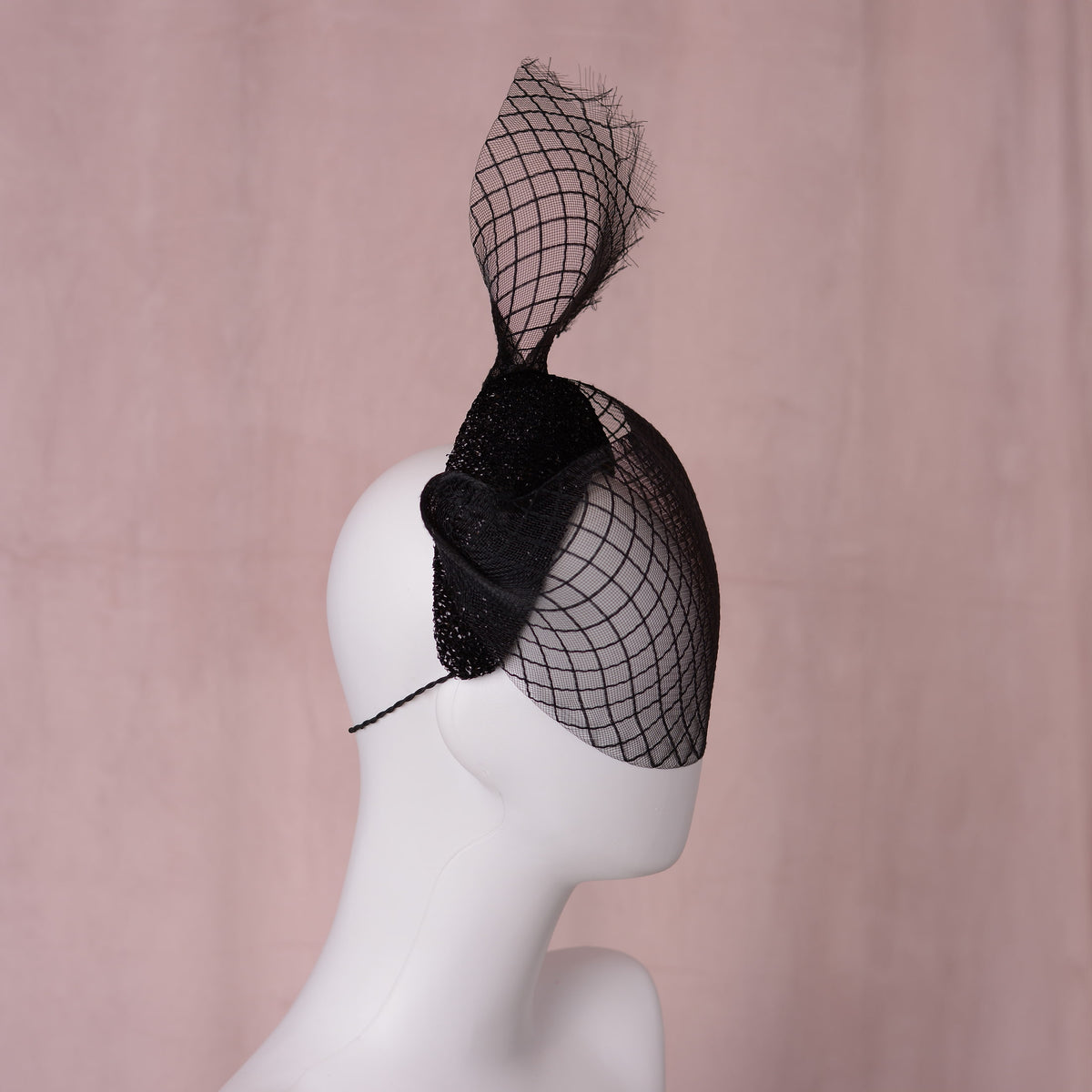 A black fascinator hat with a large sculpted crinoline veil with a diamond print pattern and a glittering headband base displayed on a white mannequin head against a soft pink background. An elegant gothic style perfect for pairing with a little black dress for a vampish look.