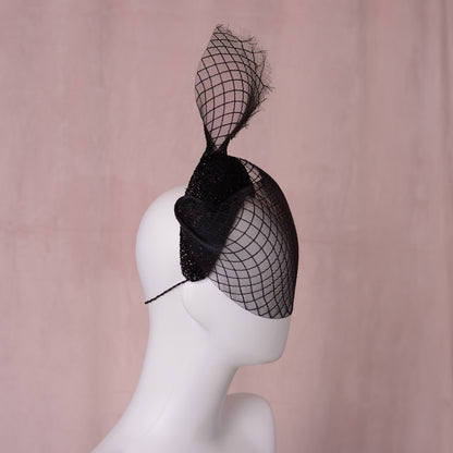 A black fascinator hat with a large sculpted crinoline veil with a diamond print pattern and a glittering headband base displayed on a white mannequin head against a soft pink background. An elegant gothic style perfect for pairing with a little black dress for a vampish look.