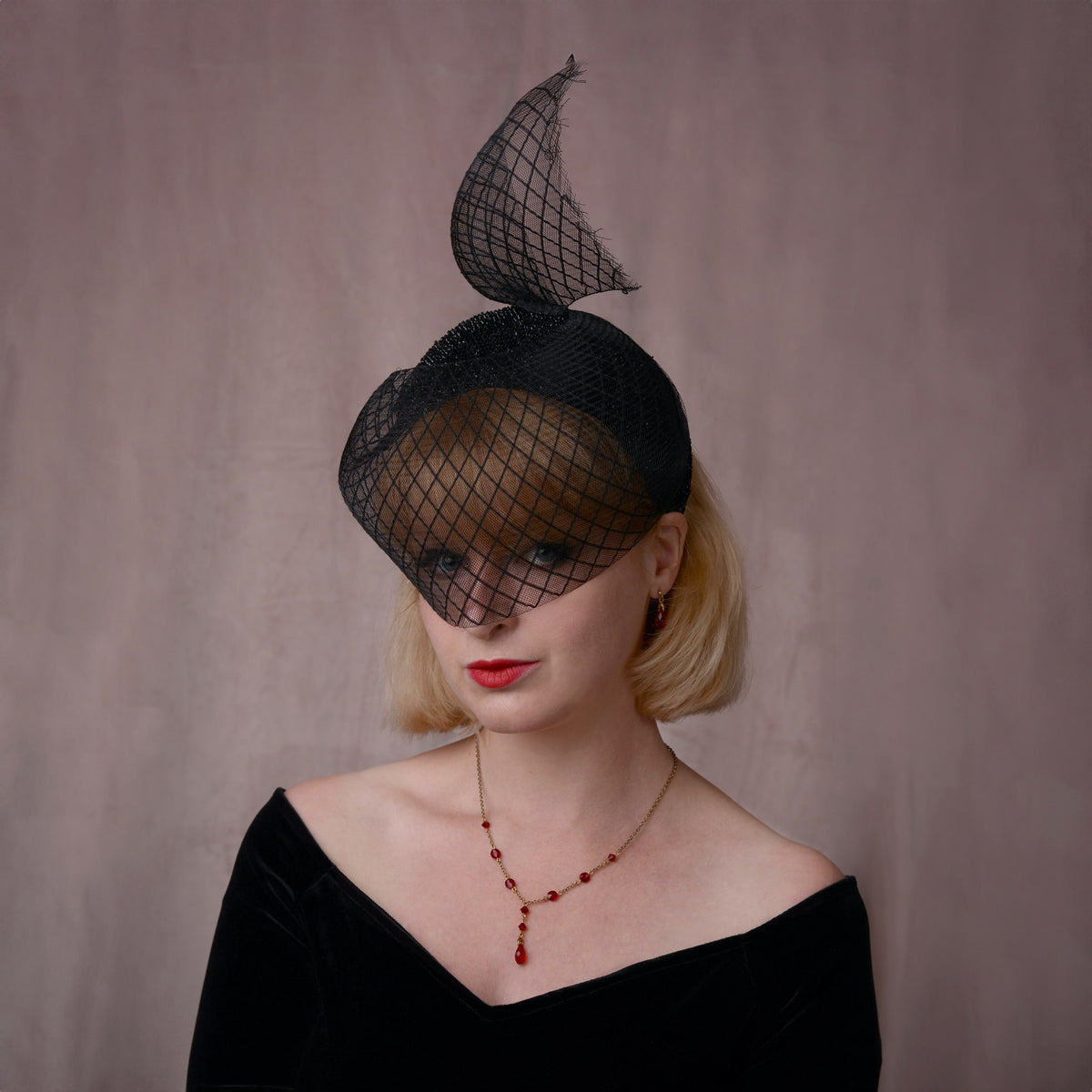 A woman with short blonde hair wears an elegant black fascinator hat with a sheer crin veil with a diamond print and a sweeping tail, paired with a black off-the-shoulder top and a red beaded necklace, set against a textured mauve background.