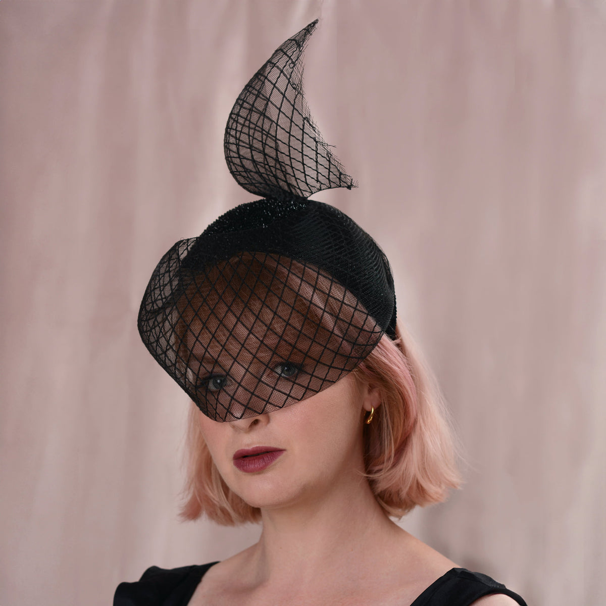 A woman with short pink hair wears a black fascinator hat with a large diamond net veil and a curved top loop, photographed against a soft beige backdrop.
