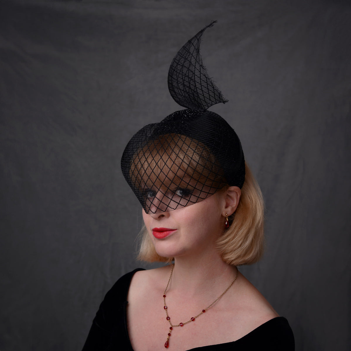 A woman with short blonde hair models a black fascinator hat with a sculpted, sweeping crin veil. She is wearing a black off-the-shoulder top and a red beaded necklace, set against a dark grey studio backdrop.