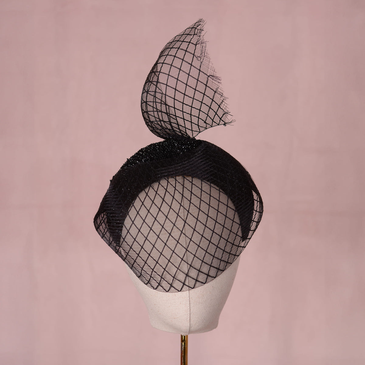 A black fascinator hat with a large sculpted crin veil with a diamond print pattern and a glittering headband base displayed on a cream-colored mannequin head against a soft pink background. An elegant gothic style perfect for pairing with a little black dress for a vampish look.