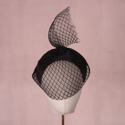 A black fascinator hat with a large sculpted crin veil with a diamond print pattern and a glittering headband base displayed on a cream-colored mannequin head against a soft pink background. An elegant gothic style perfect for pairing with a little black dress for a vampish look.