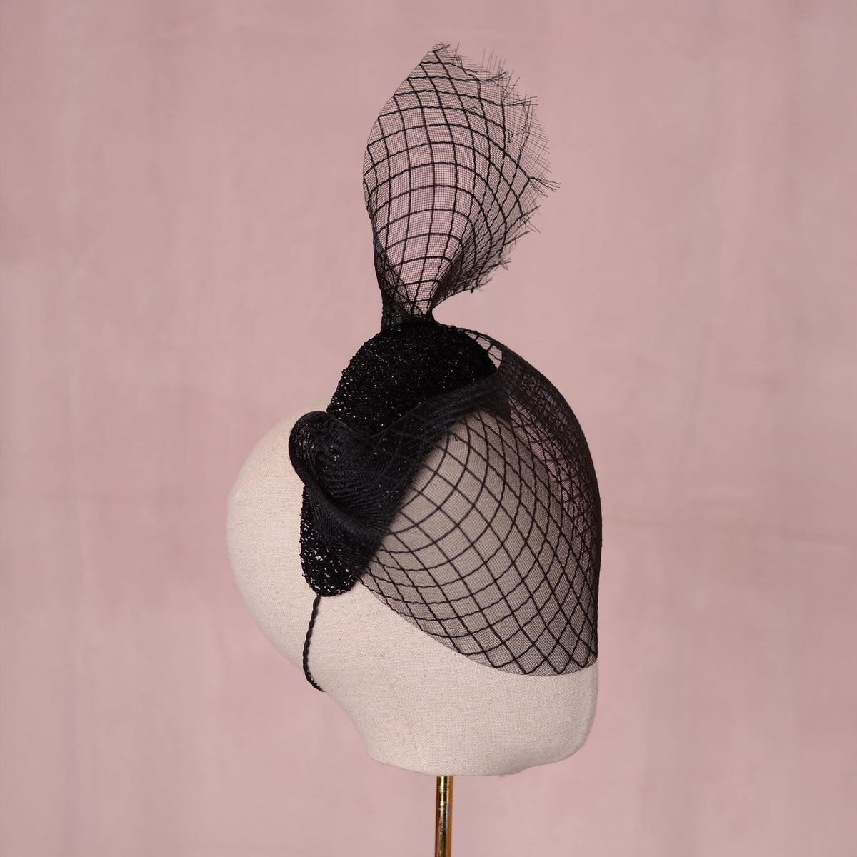 A black fascinator hat with a large sculpted crin veil with a diamond print pattern and a glittering headband base displayed on a cream-colored mannequin head against a soft pink background. An elegant gothic style perfect for pairing with a little black dress for a vampish look.