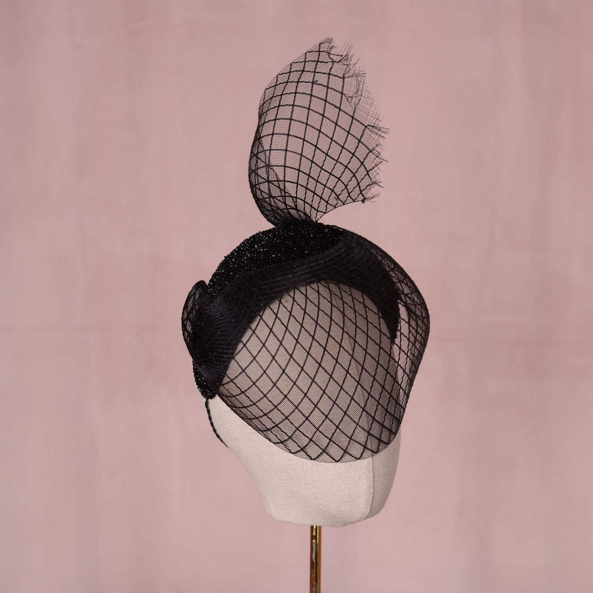 A black fascinator hat with a large diamond-patterned net veil. Sculpted into a sweeping curve with a tail above the head, displayed on a cream canvas mannequin head against a soft pink background.