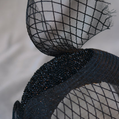 Close-up of a black headband with a decorative net overlay. The headband features a textured gliter fabric headband  and patterened net veil, creating a layered effect. The net has a diamond pattern.