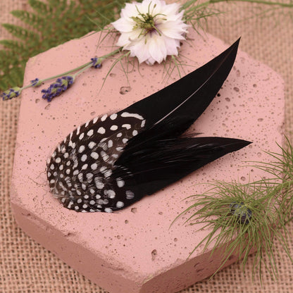 black and white polka dot feather fascinator hair clip on a pink textured stone surrounde by flowers and greenery.