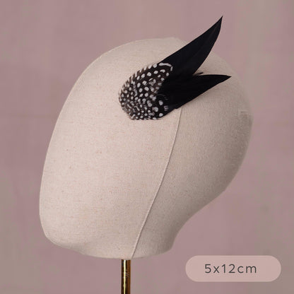 A beige fabric-covered mannequin head display stands against a dusty mauve background, wearing a black feather fascinator with a black-and-white spotted feathers. A size label reading 5x12cm appears in the lower right.