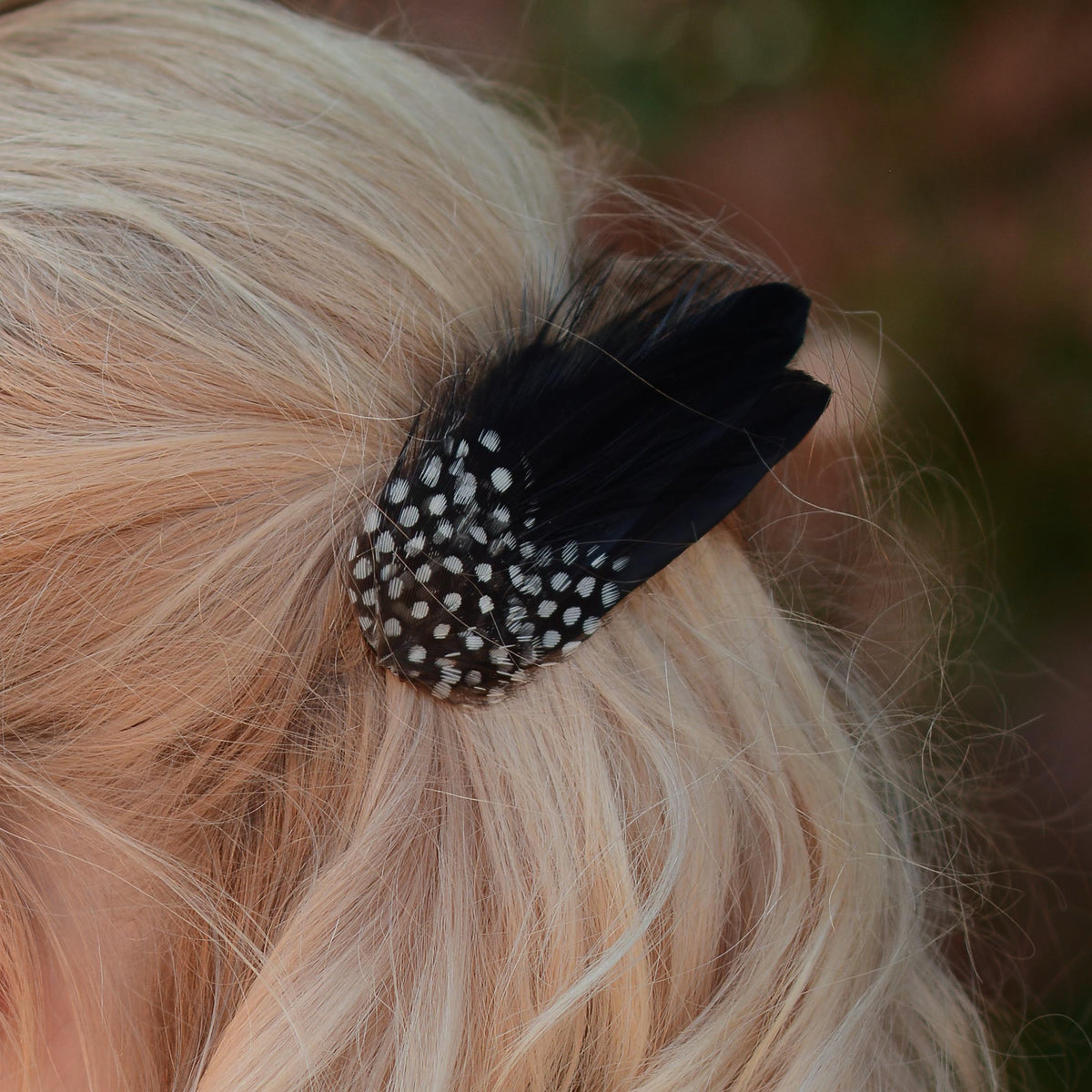 A close up of a blonde woman shown from the back wearing a black feather hair clip with white polka-dots, set against a blurred outdoor background.